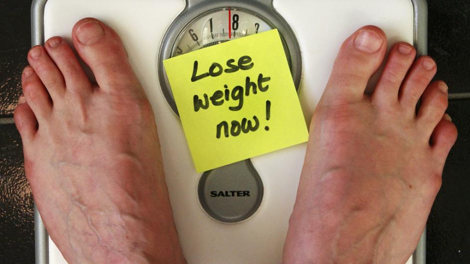 person standing on scale with sticky note that says "Lose weight now!"